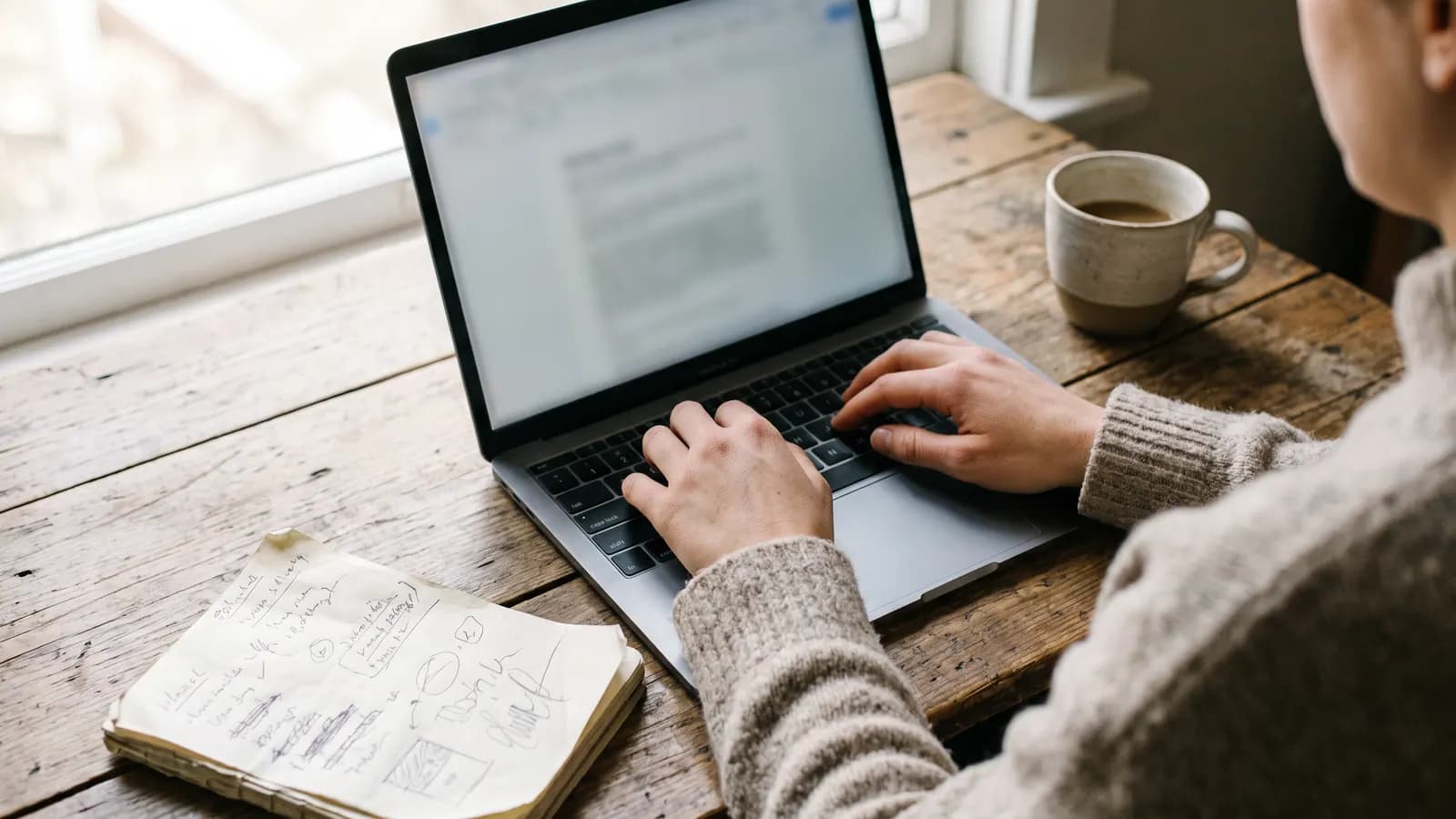 Over-the-shoulder view of someone typing on a laptop at a wooden table, with a notebook of sketches and a cup of coffee beside it