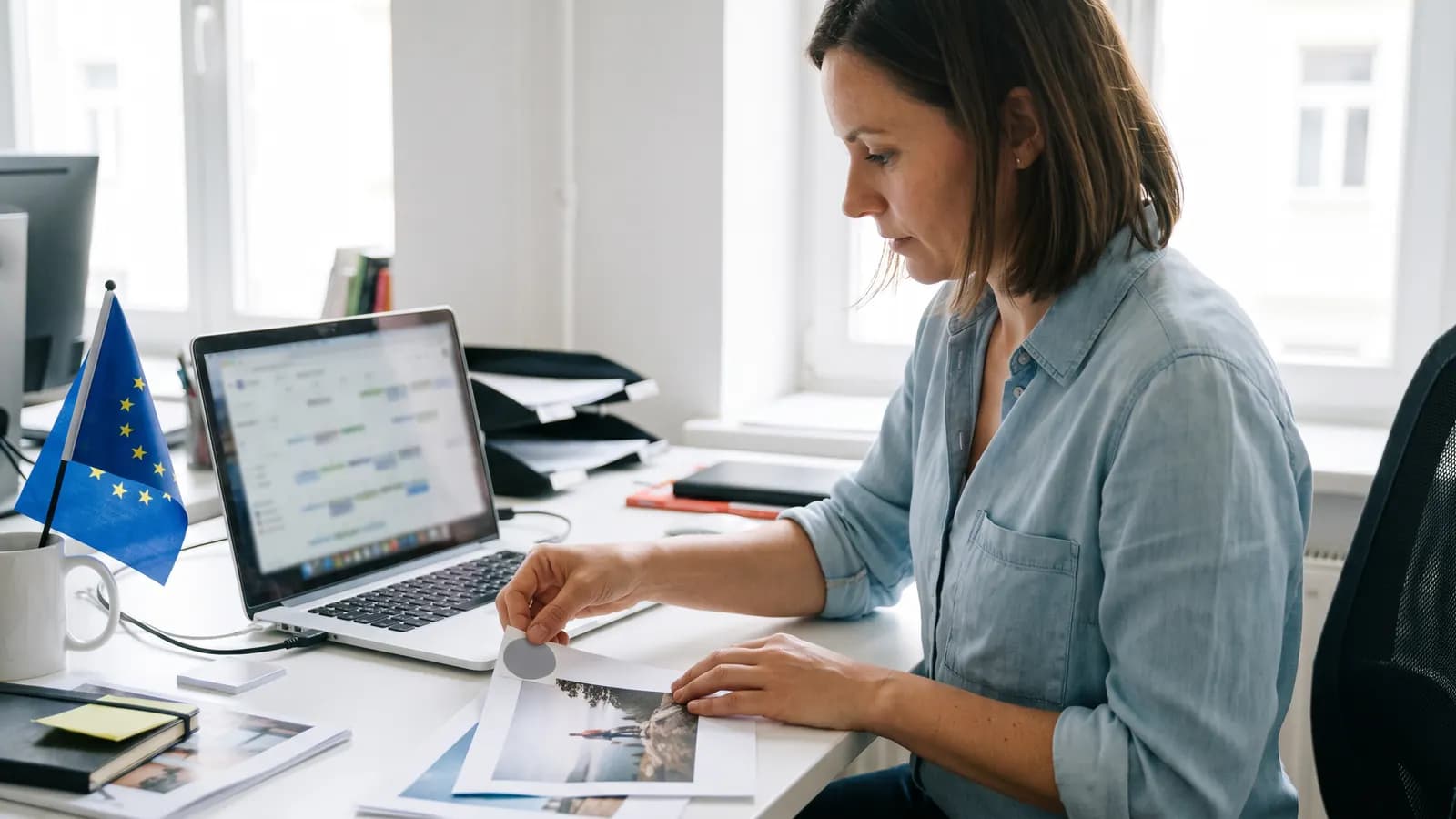 A woman at a desk sticking a small label on a printed photo, a laptop with a content calendar and a small EU flag beside her