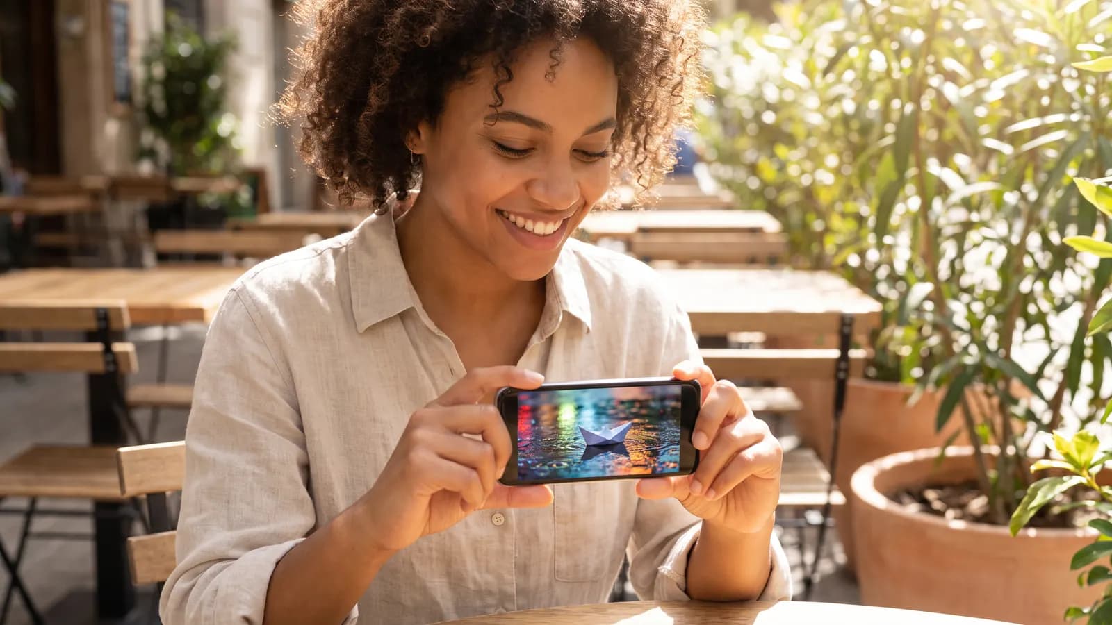 A smiling young woman at a sunny café terrace holding her phone horizontally, watching a short video of a paper boat sailing in a rain puddle